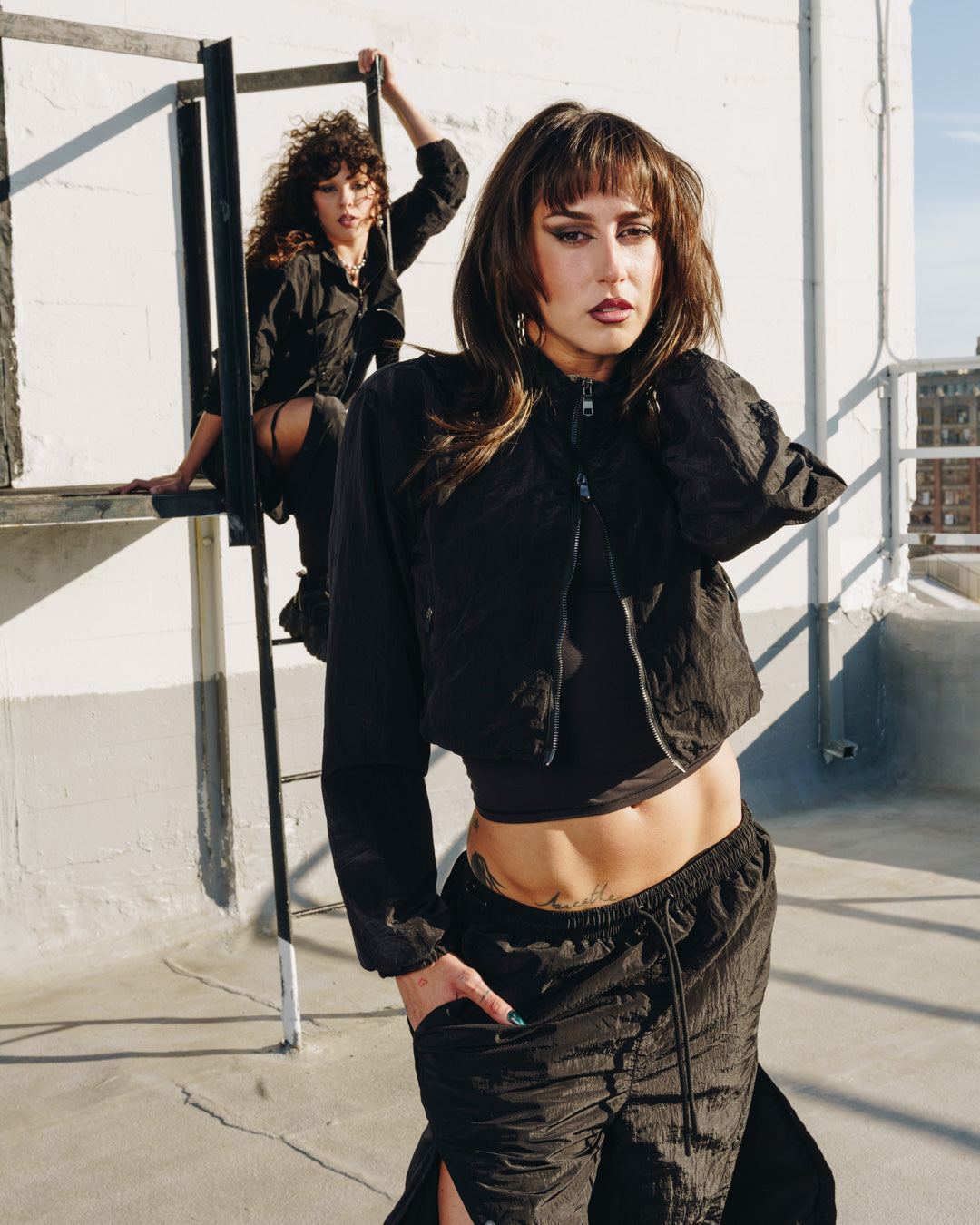 Models in the 12Twelve Off-Duty Set featuring black Track Pants and Crop Jackets, photographed on a rooftop with one model climbing and one standing.